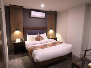 A hotel room with a bed, two bedside tables, a nightstand, an air conditioning unit, and a small table with a chair.