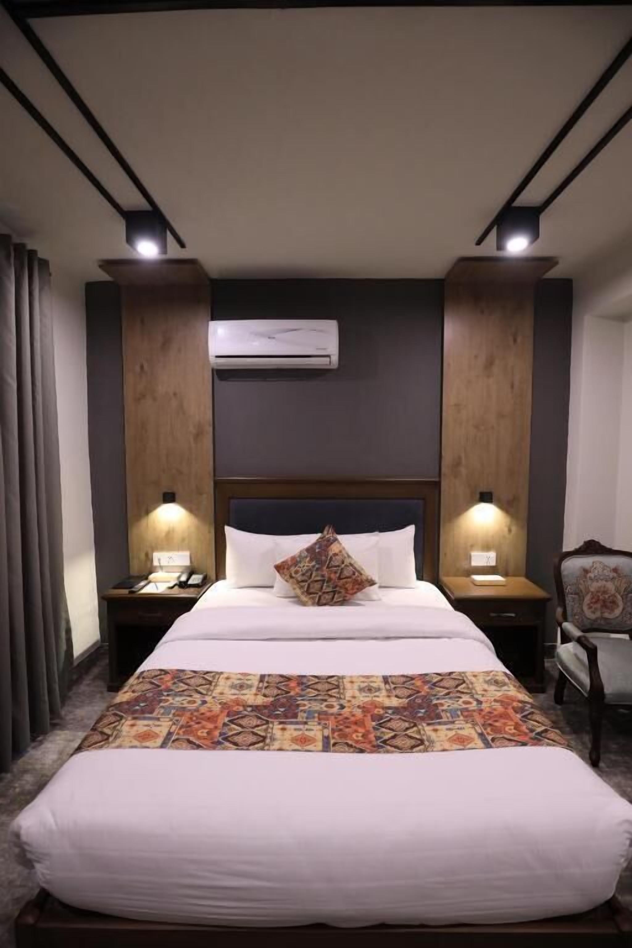 A hotel room with a large bed, two bedside tables, an air conditioning unit, and a chair.