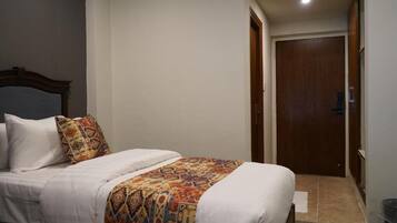 Un dormitorio con cama, almohadas, manta estampada, puerta de madera y suelo de baldosas.