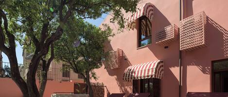 A building with a pink facade, outdoor seating with red cushions, and a large tree providing shade.
