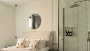 A hotel room with a bed, a round mirror, a shower, and toiletries on the wall.