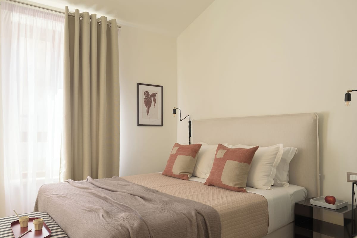 A neatly made bed with a headboard, a bedside table with a lamp, and a window with curtains.