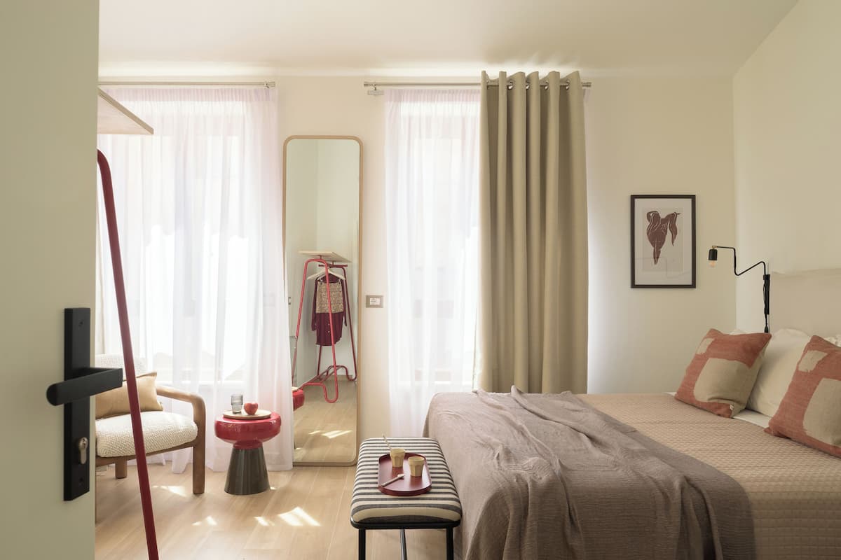 Quarto com cama, cadeira, mesinha com bandeja, espelho e porta que dá para outro cômodo.
