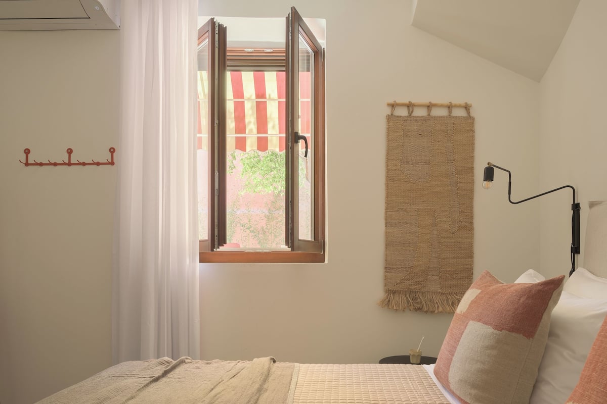 Um quarto com cama, janela com cortinas, luminária fixada na parede e tapeçaria trançada.