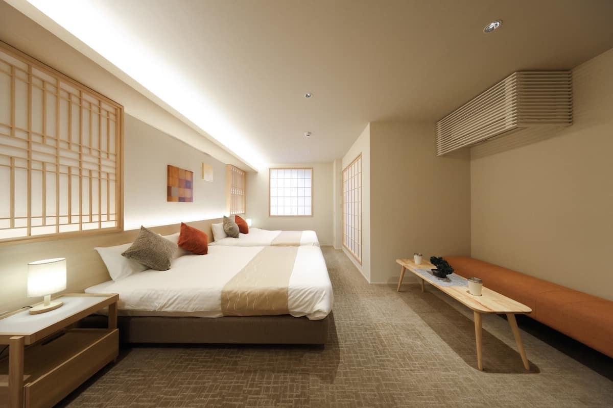 Una habitación de hotel con dos camas, una mesita de noche con lámpara, un banco y un ventanal grande.