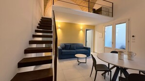 A modern interior with a staircase leading to a loft, a dining area with a round table, and a blue sofa. - Molo Mazzini (Gallipoli)