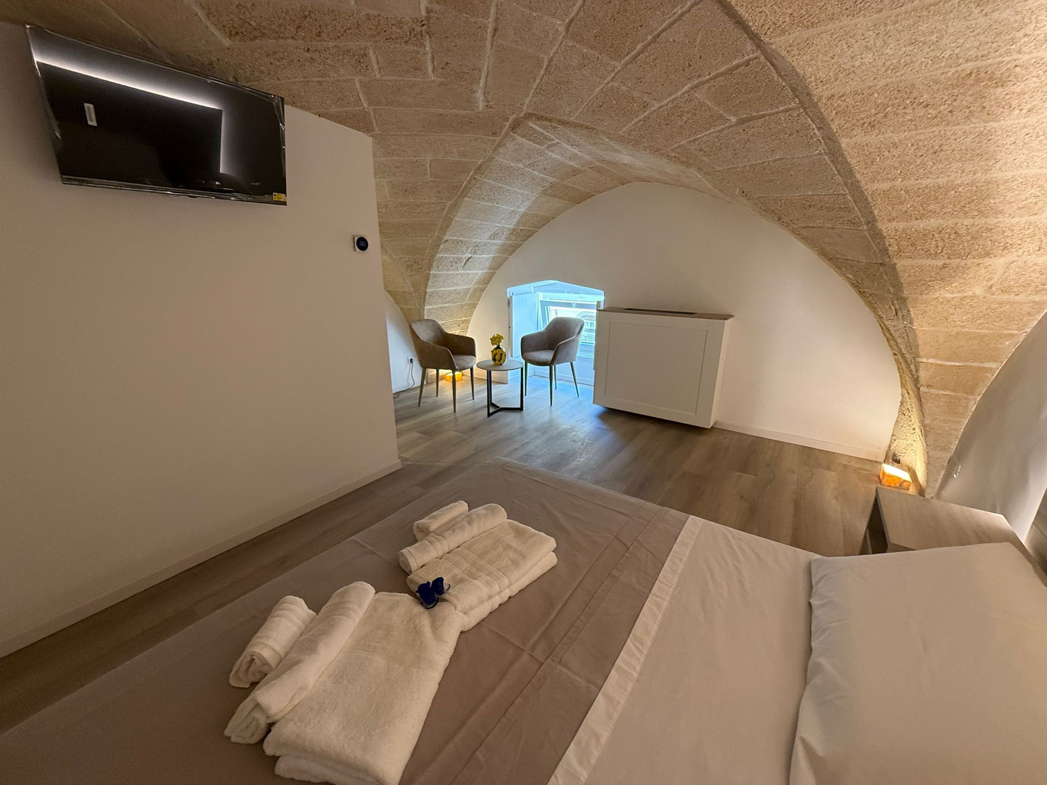 A modern room with a stone ceiling, a bed with white linens, a flat-screen TV, and a small seating area with chairs and a table.