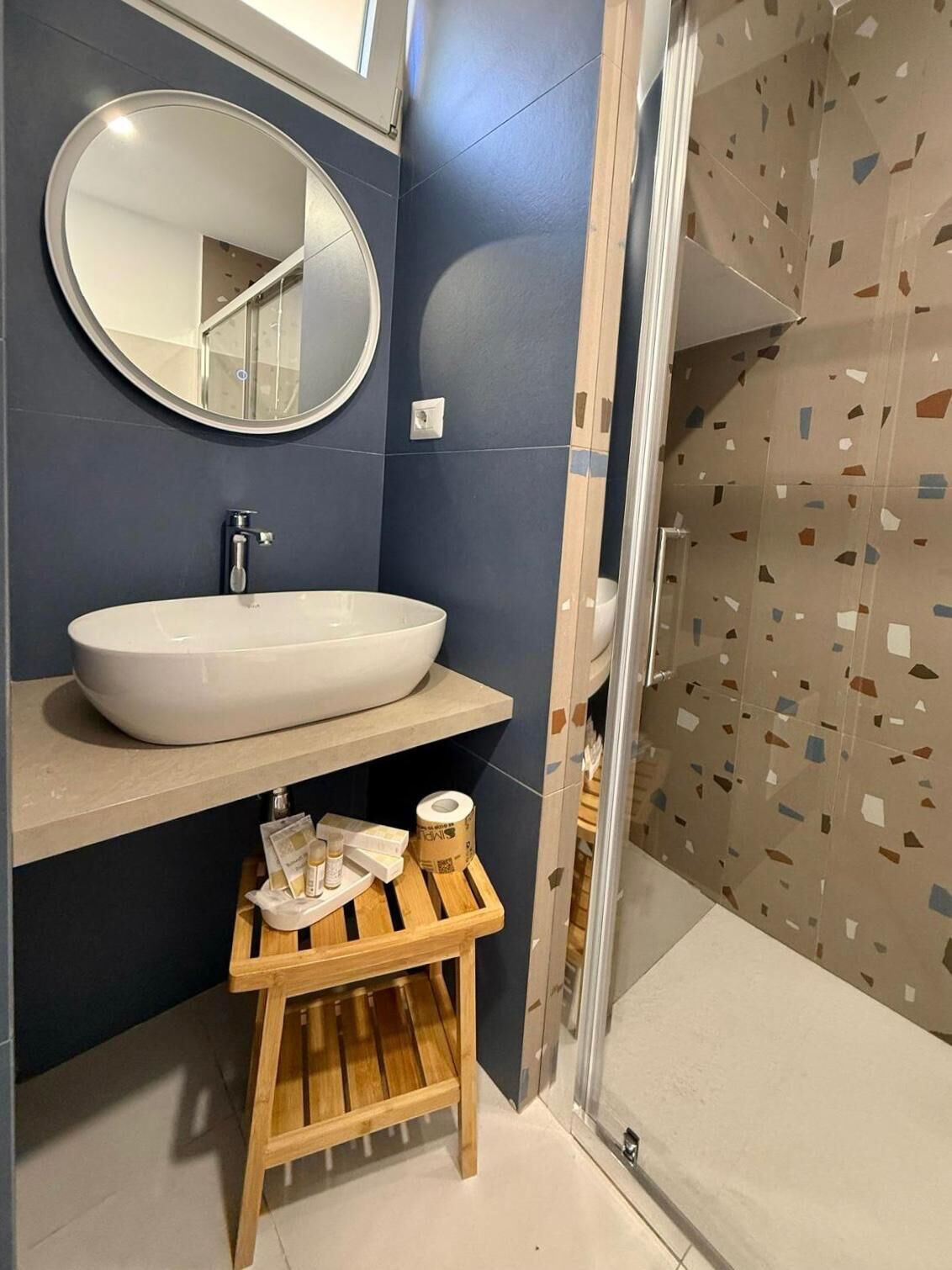 A bathroom with a large oval sink, a round mirror, a wooden stool with toiletries, and a shower with a geometric tile pattern.