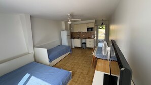 Una habitación compacta con dos camas individuales, una kitchenette pequeña, una mesa de comedor y un ventilador de techo.