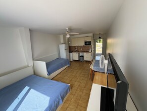 Ein kompakter Raum mit zwei Einzelbetten, einer kleinen Küchenzeile, einem Esstisch und einem Ventilator, sowie einem Deckenventilator.