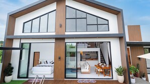 Una casa moderna de dos pisos con un amplio acceso por puerta de vidrio, un dormitorio visible con cama y sofá, un comedor con mesa y sillas, y una escalera que sube al piso superior.