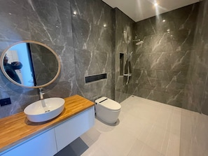 Een moderne badkamer met een marmeren wand, een ronde spiegel, een houten wastafelmeubel, een witte wasbak, een toilet en een douchecabine met glazen wanden.
