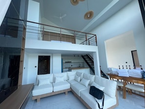 Een modern interieur met een witte modulaire bank, een eettafel met opgevouwen beddengoed, een keuken en een trap naar een hoger gelegen verdieping.
