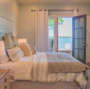 Un dormitorio con cama, almohadas, una mesita de noche, una lámpara y vistas al exterior a través de una puerta.