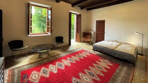Quarto com uma cama, um tapete estampado vermelho, uma cadeira, uma mesa pequena e uma janela com vista para vegetação.