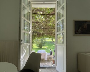 An open door leading to a garden with a table and chairs, a person in the distance, and a painting on the wall.