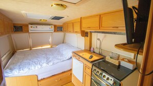 Interior de una caravana pequeña con cama, kitchenette y un refrigerador compacto.