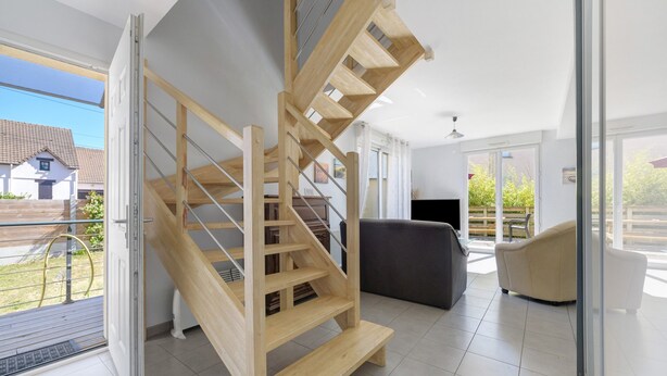 A modern interior with a wooden spiral staircase, a grey sofa, and a large window offering a view of the outside. - Gîte des Pluviers – Modern Villa Near the Beach in Saint-Martin-de-Bréhal (Bréhal)