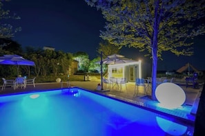 A swimming pool with a small gazebo and chairs, surrounded by trees and a well-lit patio area.