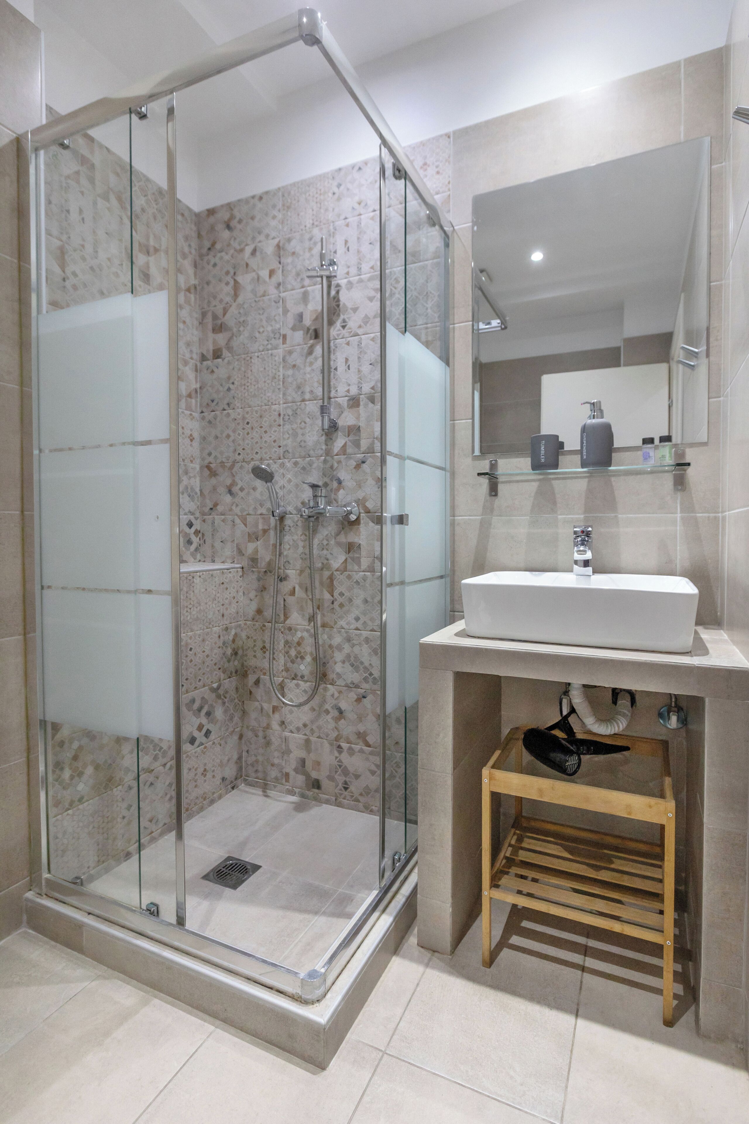 A modern bathroom with a glass-enclosed shower, a sink with a mirror above, and a wooden shelf.
