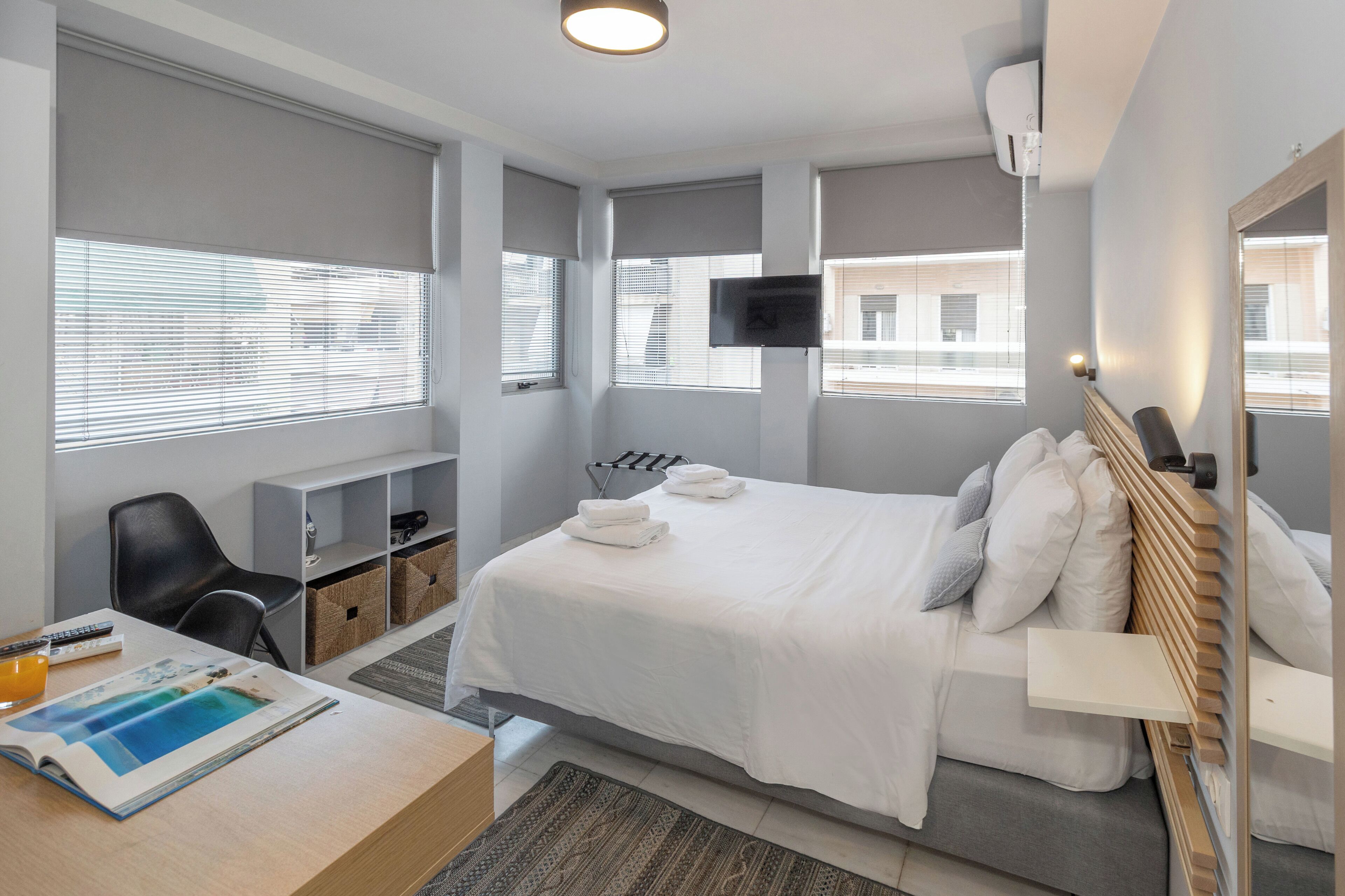 Een moderne hotelkamer met een bed, een bureau met een tijdschrift, een stoel, een televisie en een raam met jaloezieën.