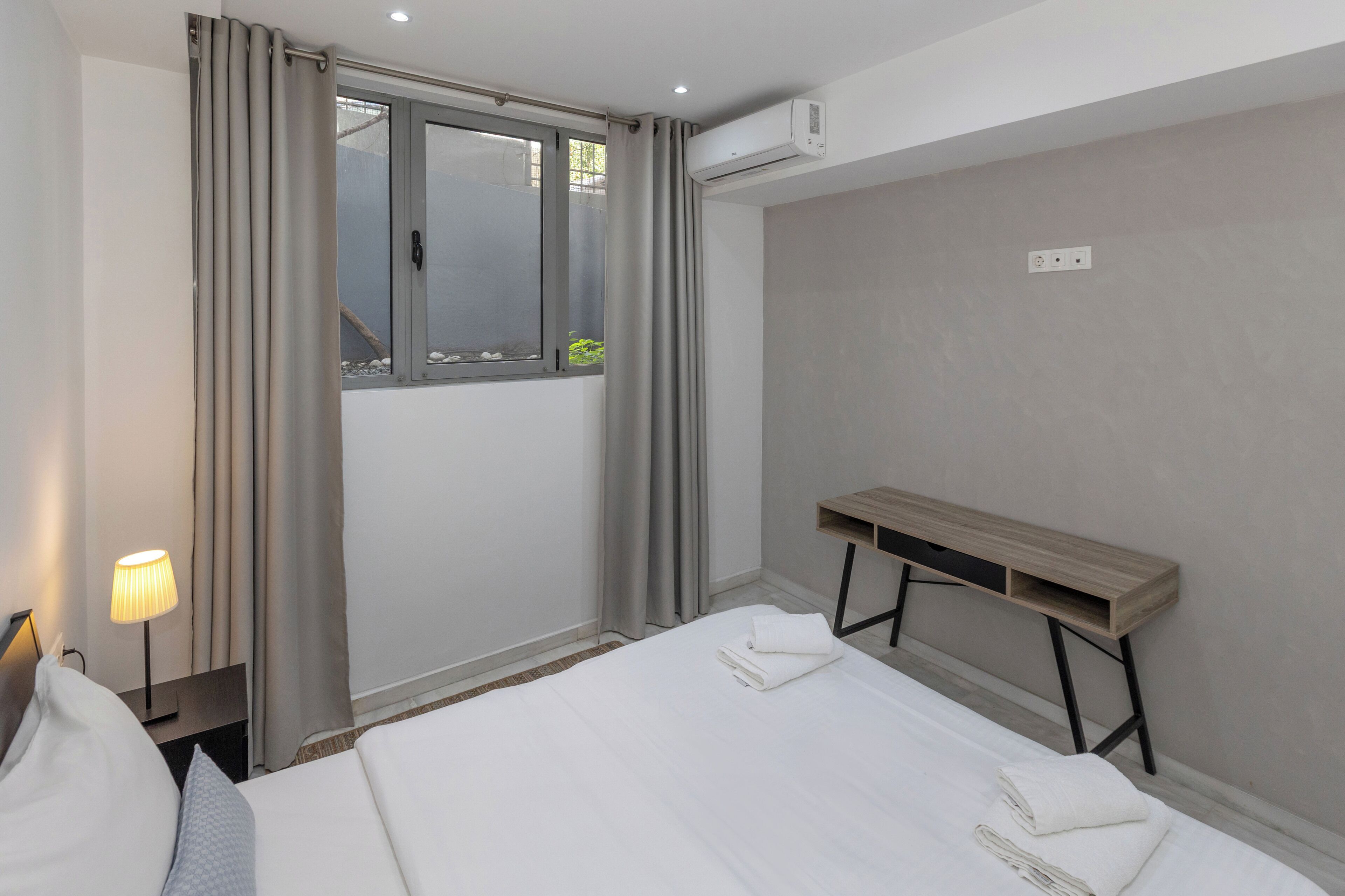 A hotel room with a bed, a desk, a lamp, a window with curtains, and an air conditioning unit.