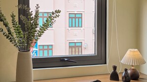 Een bureau met stoel, een raam met uitzicht op gebouwen, een lamp en een vaas met planten.