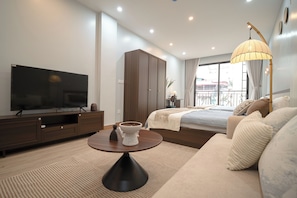 Una habitación de hotel moderna con televisor de pantalla plana, armario de madera, mesa de centro redonda, sofá y balcón con cortinas.
