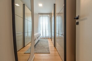 A modern hotel room with a large mirror, a bed with a white headboard, a wooden floor, and a view of the city through large windows. - Zaria Luxury Residence (Zadar)