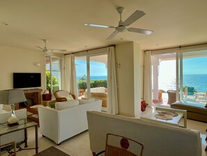 Ein geräumiges Wohnzimmer mit Fernseher, weißen Möbeln, Deckenventilator und großen Fenstern mit Meerblick.