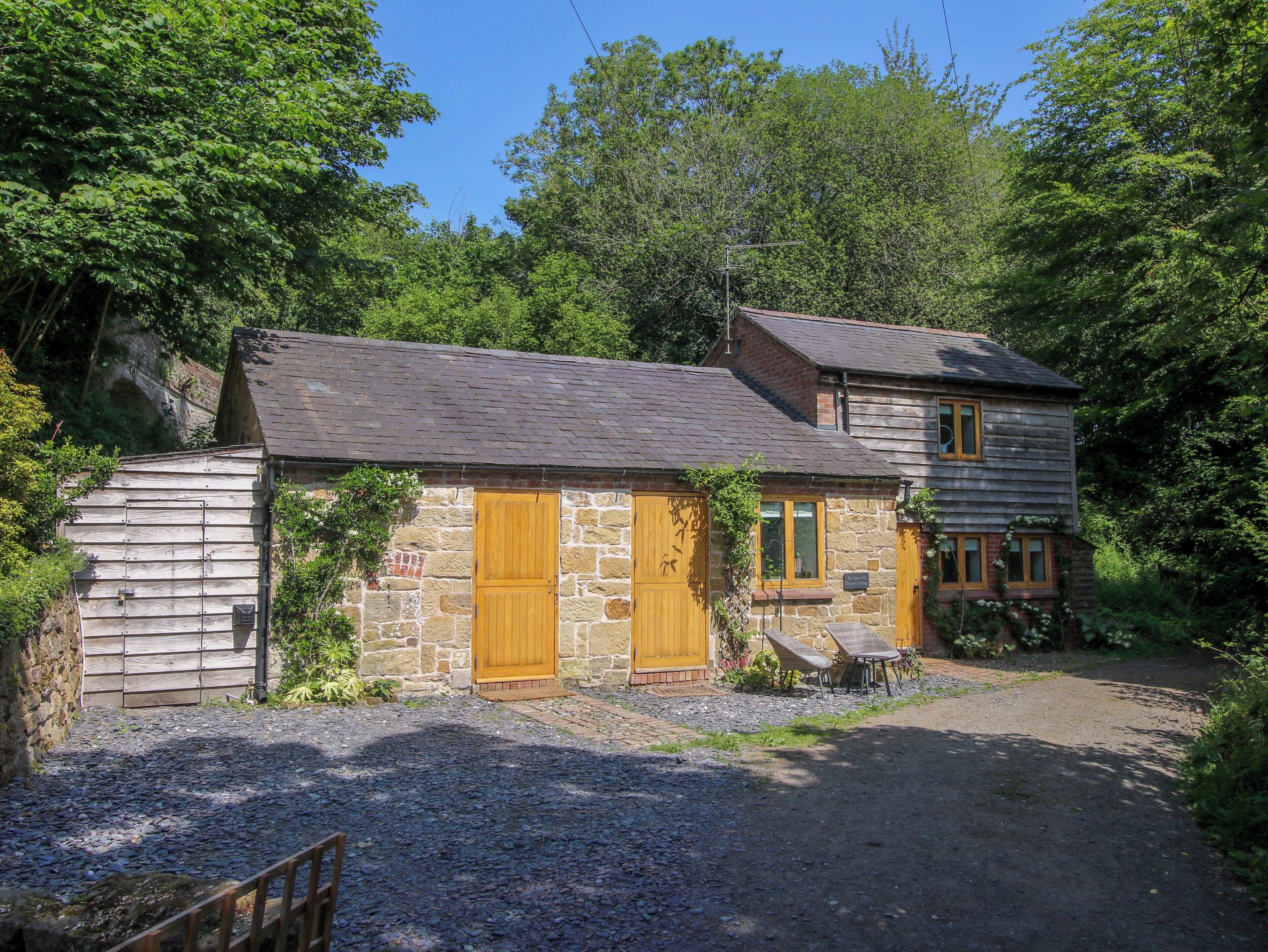 A rustic stone building with a wooden door and a small porch with two wicker chairs.