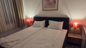 Ein ordentlich bezogenes Bett mit weißen Leinen, zwei Nachttische mit Lampen, ein gerahmtes Blumenbild an der Wand und ein Fenster mit Vorhängen.