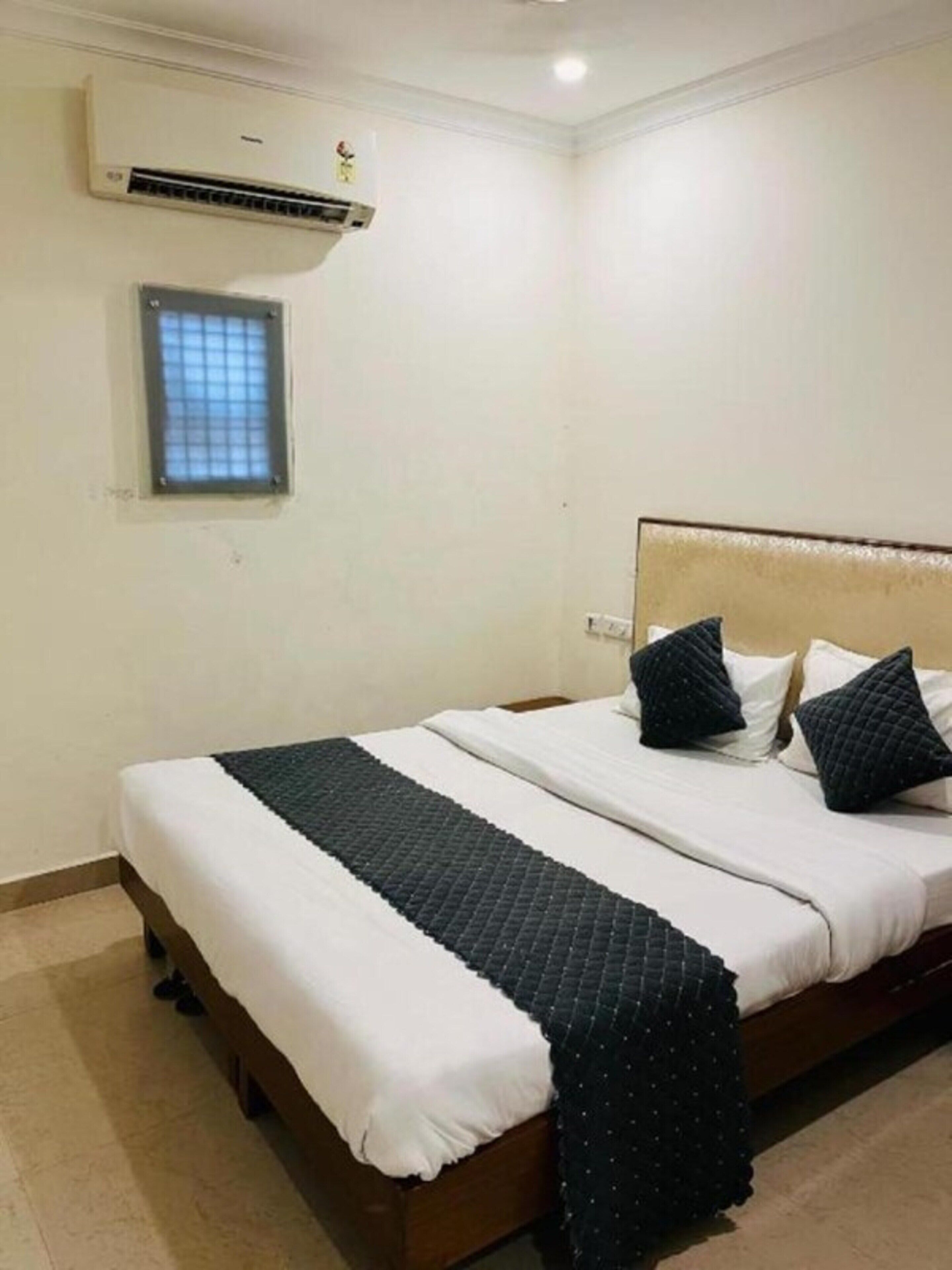 A neatly made bed with white and dark bedding, a small window with a grid pattern, and an air conditioning unit on the wall.