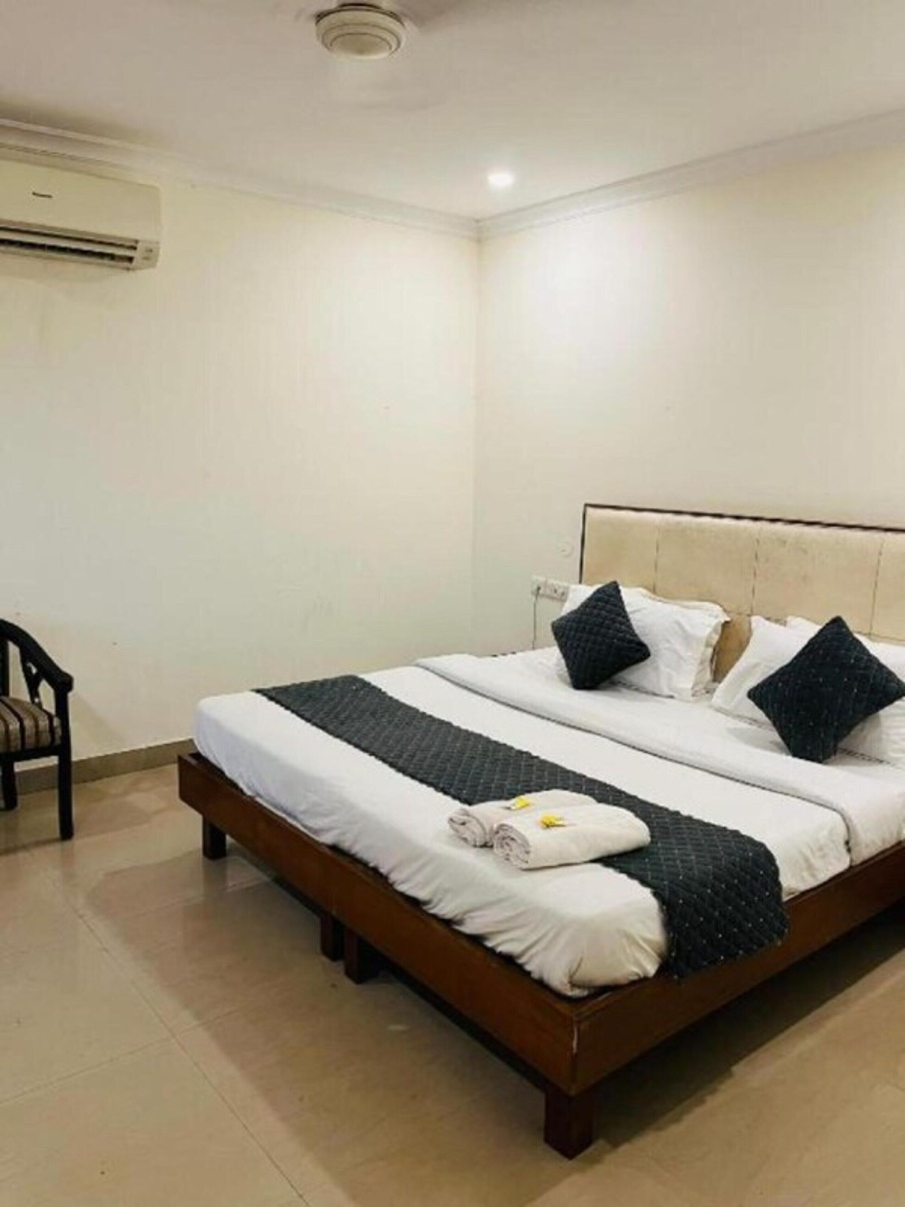 A neatly made bed with pillows, a folded towel, and a chair in a simple room with a tiled floor and an air conditioning unit on the wall.