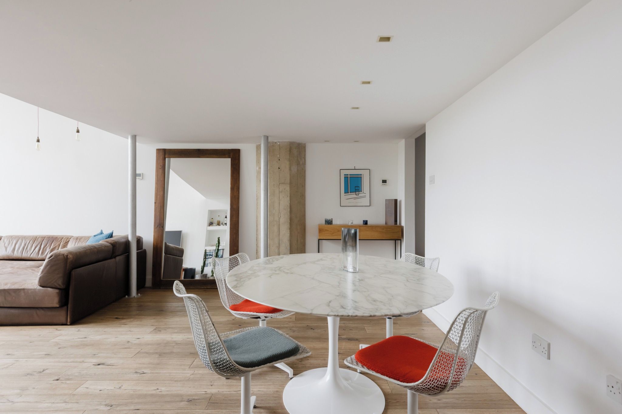 Een moderne woonkamer met een ronde marmeren tafel en stoelen van draadstaal, een bruine bank en een houten vloer.