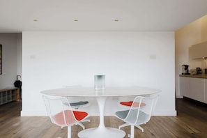 A minimalist dining area with a round table and two chairs, a white wall, and a kitchen area in the background. - Sunny 1BD Loft Style Flat in the Heart of Hackney (London)