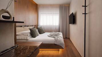 Una habitación de hotel moderna con cama, piso de madera, mesita de noche de mármol, televisor montado y ventana con cortinas translúcidas.
