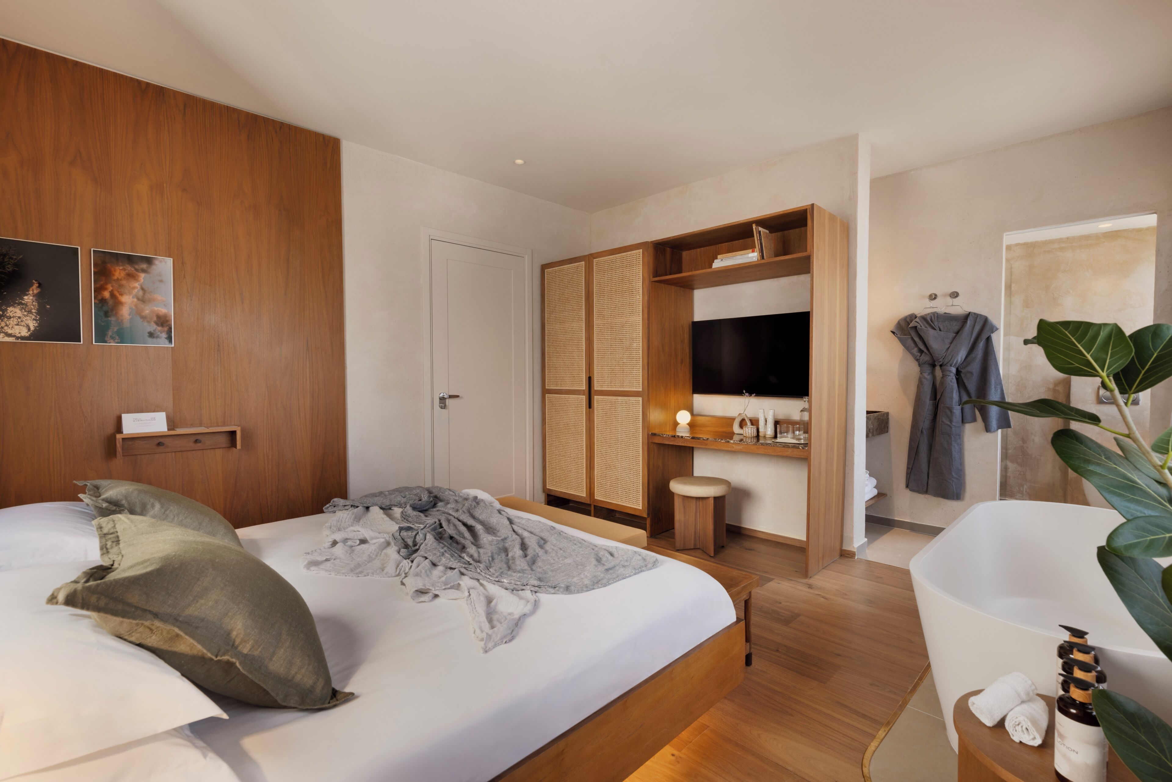 A modern bedroom with a large bed, a wooden wardrobe, a TV, a bathroom with a bathtub, and a plant.