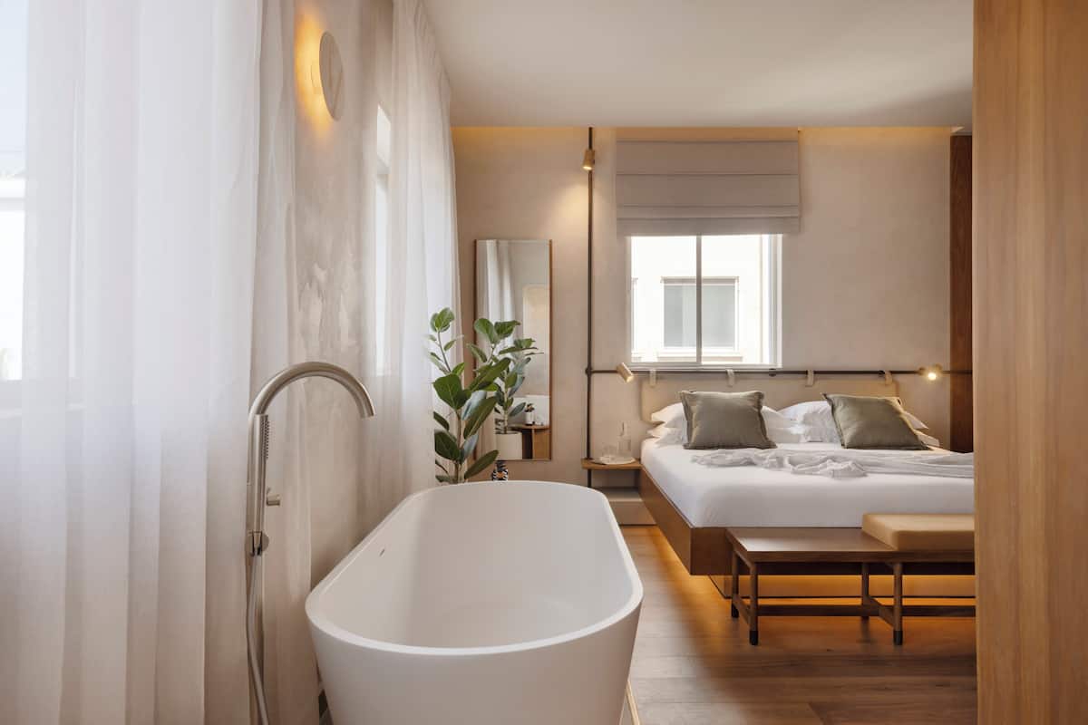 A modern bedroom with a large bathtub, a bed with pillows, a bedside table, and a window with a view.