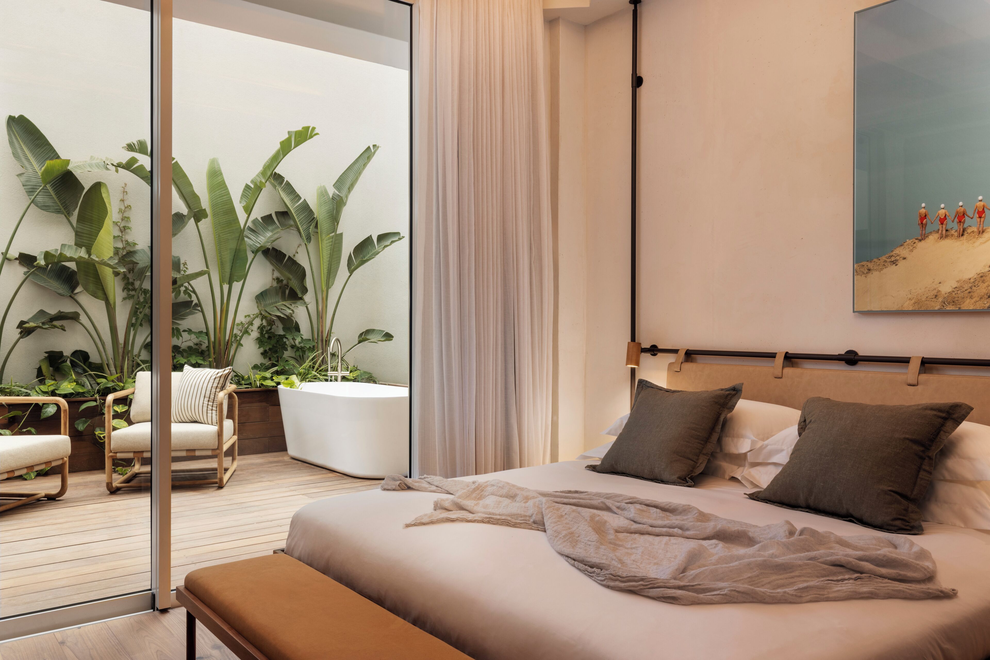 Un dormitorio con una cama grande, un banco, una planta en maceta, una silla y vistas a un balcón con plantas.
