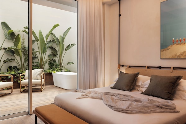A bedroom with a large bed, a bench, a potted plant, a chair, and a view of a balcony with plants. - The ABY Hotel (Tel Aviv)