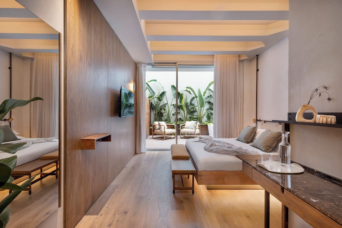 A modern hotel room with a wooden floor, a sofa, a TV, a wooden shelf, and a view of a balcony with plants.