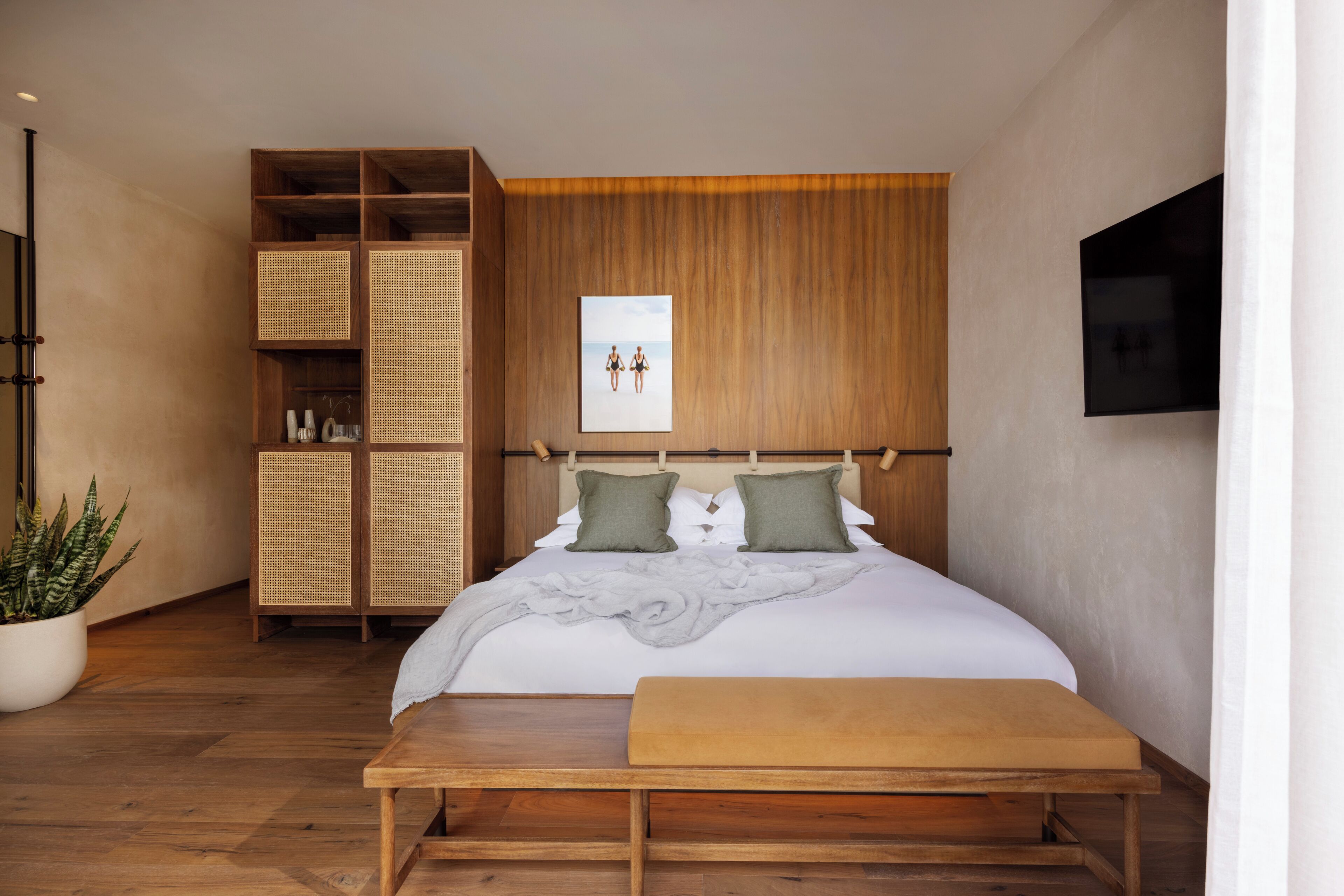 A bedroom with a wooden headboard, a bed with green pillows, a bedside table, a large potted plant, and a wall-mounted TV.