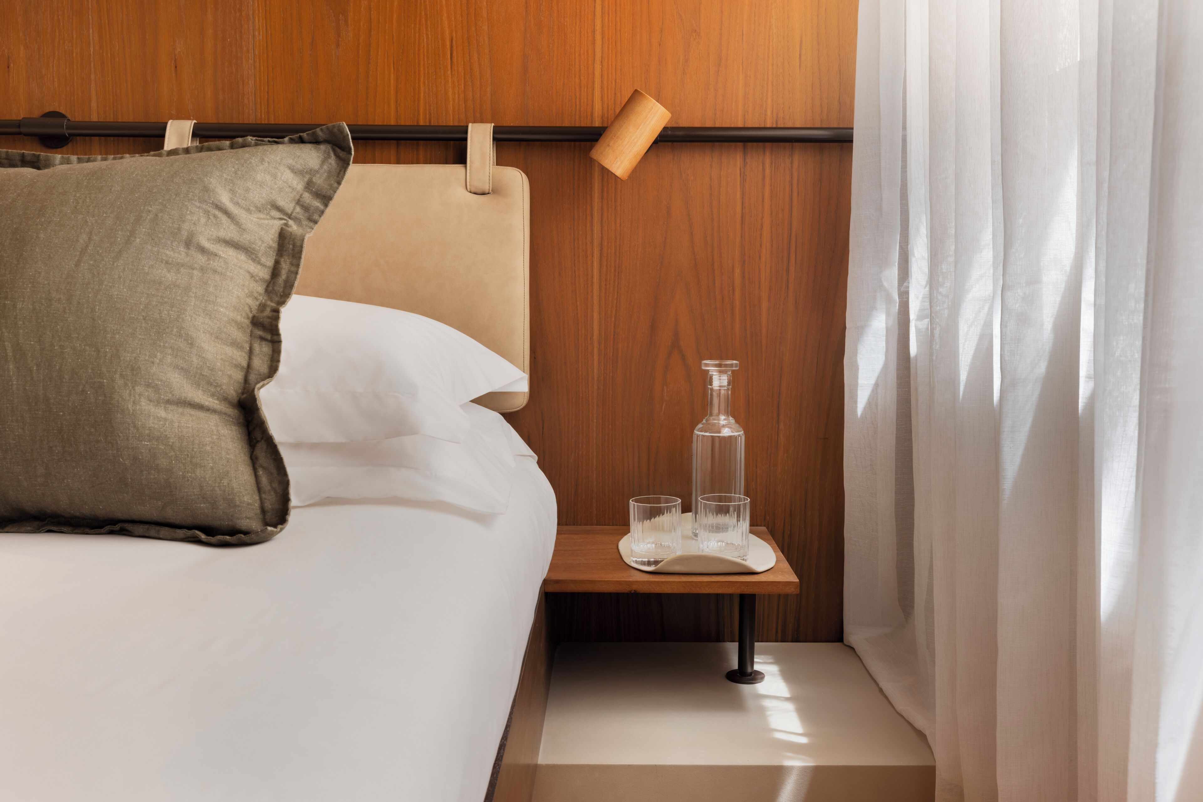 A neatly made bed with pillows, a bedside table with glasses and a bottle, and a wooden headboard.