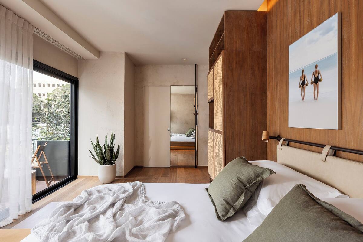 A bedroom with a bed, a potted plant, a wooden headboard, and a view through a window.