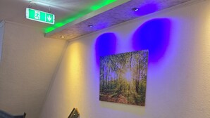 A hallway with a green carpet, a staircase, a wall-mounted emergency exit sign, and a forest-themed painting. - Gästehaus Bodewurm (Braunlage)