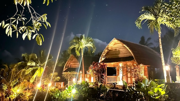Dos cabañas de madera con techos de paja, rodeadas de vegetación tropical e iluminadas con luz ambiental.