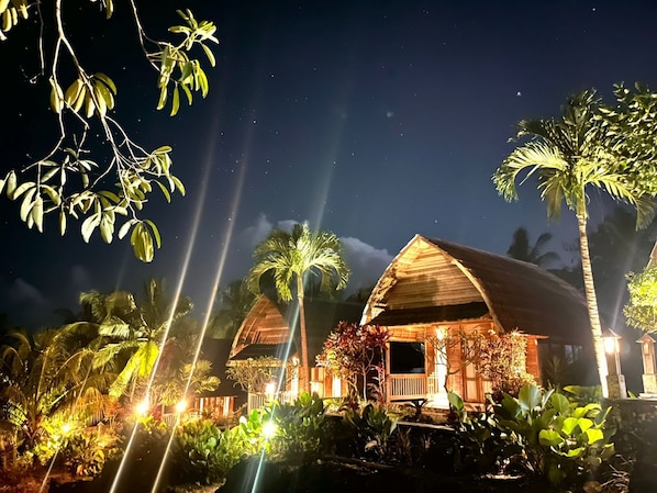Two wooden cabins with thatched roofs, surrounded by tropical vegetation and illuminated by ambient lighting.