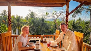Dos personas sentadas en una mesa de madera con tazas y un bol, rodeadas de follaje tropical.