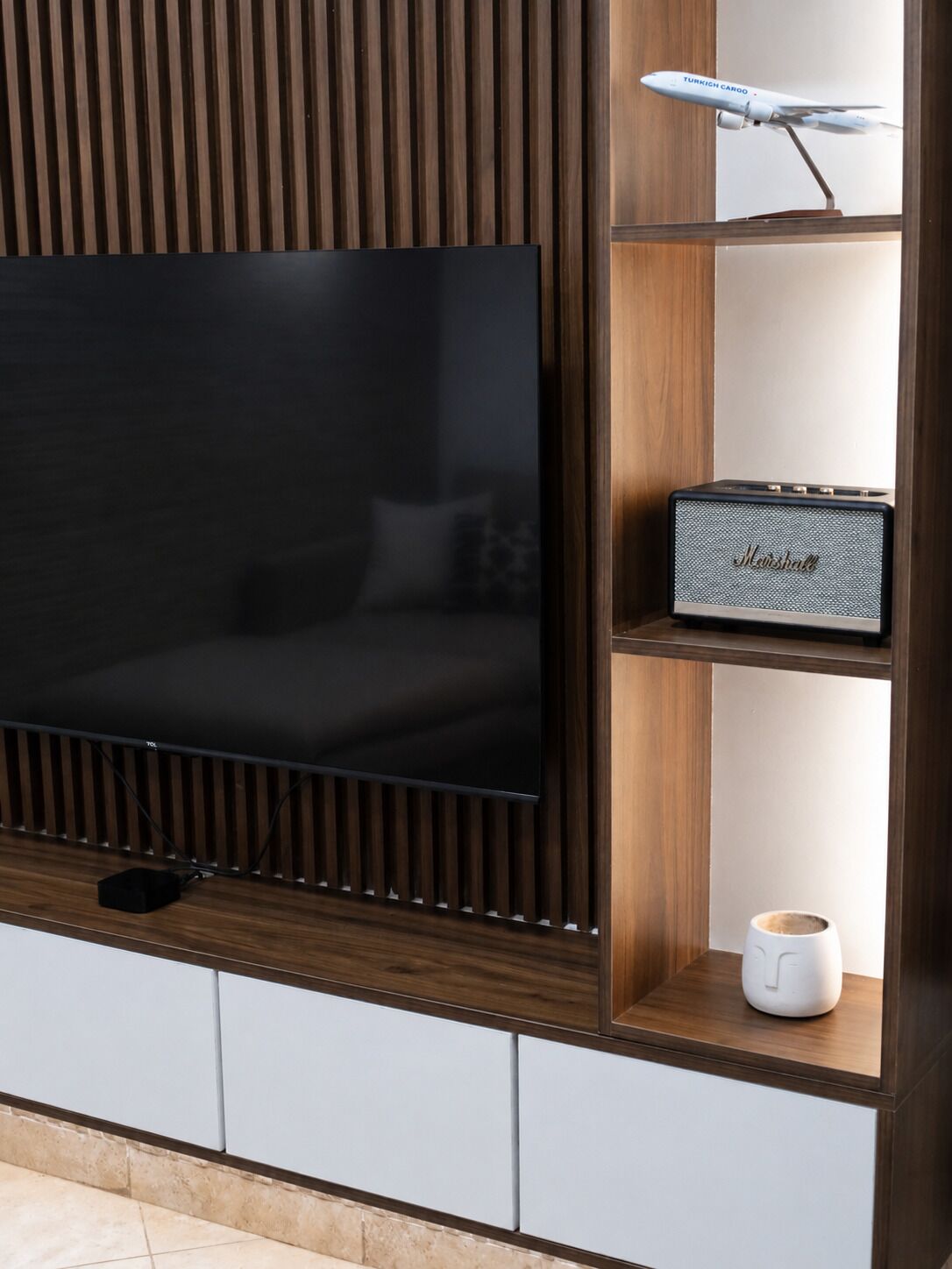 Un salon moderne avec un téléviseur à écran plat fixé sur un panneau en bois, un haut-parleur Marshall sur une étagère et une machine à café sur l’étagère inférieure.